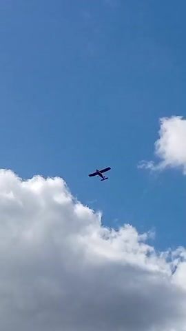 A small, dark-colored airplane with two wings and a tail is seen flying in a bright blue sky filled with white, puffy clouds. The plane is centrally located in the frame and appears to be traveling from left to right. The camera is angled upwards, capturing the underside of the airplane. The lighting suggests it is daytime, with the sun illuminating the clouds.