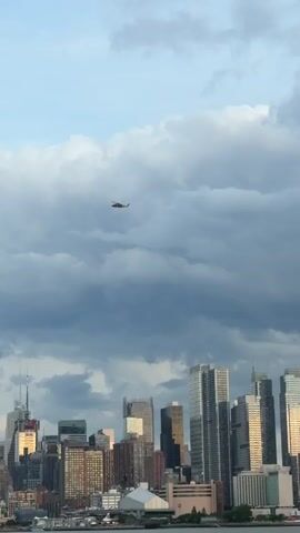 A wide shot captures a city skyline under a cloudy sky. A small helicopter flies in the upper portion of the frame. The buildings in the distance are tall and modern.