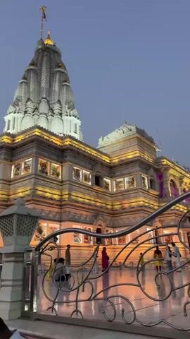 The video shows a temple with a tall, gray spire, illuminated with bright white and yellow lights. The building is decorated with many rectangular windows that are also illuminated. A fence with white posts runs along the perimeter in front of the temple. People are walking by the fence. The color of the temple lights changes from yellow to green. The sky above the temple is a muted blue.