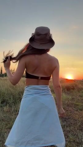 A woman stands in a field wearing a cowboy hat, a bikini top, and a light blue skirt tied around her waist. She has long, dark hair. The sun is setting behind her, casting a warm glow over the scene. The woman is facing away from the camera, with her head turned slightly to her left. She is touching her hair. The field is covered in dry grass and some scattered bushes. The sky is a gradient of orange, pink, and blue. The overall impression is one of a serene and beautiful natural setting.