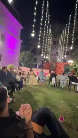 The video shows a nighttime outdoor event, likely a wedding reception, taking place on a grassy lawn. People are seated in chairs, facing a brightly colored stage-like structure adorned with striped fabric in red, yellow, and blue. String lights hang above the lawn, creating a festive atmosphere. The foreground shows a person lying on the grass, their legs visible in the frame. The people in the chairs include men, women, and children, dressed in a variety of formal and semi-formal attire. Some people are watching the stage.