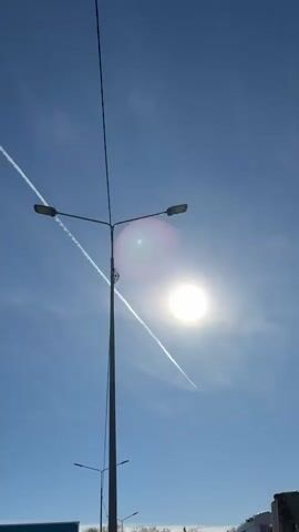 The video shows a bright blue sky with two intersecting contrails created by high-flying aircraft. The sun is visible, creating a glare, partially obscured by a tall street light. The camera pans upwards, following one of the contrails as it stretches across the sky. The end of the contrail tapers off as the camera continues to follow it. A smaller street light is seen briefly in the lower left corner.