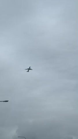 The video depicts a large airplane ascending into a cloudy, gray sky. The plane is seen from below, and the view includes glimpses of streetlights in the foreground. The camera angle is tilted upwards, tracking the plane's movement.