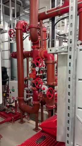 A video shows a close-up view of a red industrial pipe system with several valves and gauges. The pipes are arranged in a complex network, with some vertical and others horizontal. Red-handled valves are visible at various points, suggesting manual control over the flow of a substance. Several circular gauges are attached to the pipes, likely indicating pressure or flow rate. The surrounding environment appears to be a mechanical room or utility area, with metal framing and electrical conduits visible in the background. A white control box with a green light is mounted on a vertical pipe on the left side of the frame. The overall impression is one of a functional, industrial plumbing system.