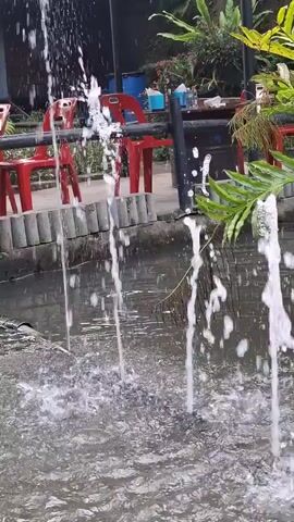 The video shows a close-up view of a small pond or water feature. In the foreground, water is spraying upwards from multiple spouts, creating a dynamic visual effect. The water is clear and reflects the surrounding environment. In the background, there is a variety of lush green plants, including ferns and other tropical foliage. Red chairs and a table are visible, suggesting an outdoor seating area. The overall impression is one of a serene and natural setting, possibly a garden or a restaurant with an outdoor patio. The video is shot at an angle that emphasizes the movement of the water and the vibrant greenery.