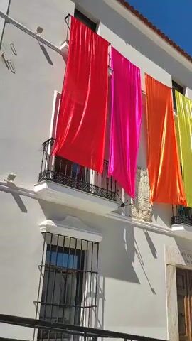The video shows a building with several colorful banners hanging from its windows. The banners are arranged in a rainbow pattern, starting with red, then pink, orange, yellow, green, blue, and purple. The building is white with a tiled roof and has balconies with black railings. There is a blue sky visible in the background. The video pans from left to right, showing the full length of the banners. The sun is shining brightly, casting shadows on the building.