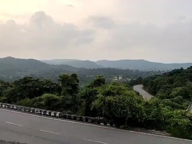 The video shows a scenic view of a winding road through a lush green landscape with rolling hills under a cloudy sky. The road is paved and has white lines marking the lanes. On the left side of the road, there is a concrete barrier with black and white patterns. Dense green foliage, including trees and bushes, borders the road, creating a tunnel-like effect in some parts. In the distance, more hills are visible, covered in trees, and hints of buildings can be seen nestled within the landscape. The overall impression is one of a peaceful, natural environment with a road that invites exploration.