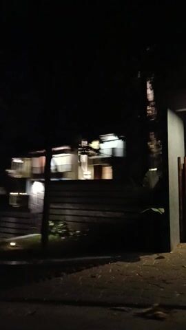 The video shows a multi-story building at night. The building has several windows, many of which are illuminated, suggesting that people are inside. The exterior of the building features a modern design with horizontal slats and balconies. Outside the building, there are trees and plants, illuminated by outdoor lighting. A fence or gate is visible on the right side of the frame. The overall scene appears to be a residential or commercial property in a dark, outdoor setting. The time of day is clearly night due to the darkness and the artificial lighting. There are no discernible people or specific activities taking place. The location context indicates Sambirata, Indonesia.