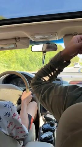 The video shows the interior of a car from the passenger's perspective, looking forward through the windshield. The driver's hands are visible on the steering wheel, and they are wearing a white shirt with a bicycle pattern and shorts. Another passenger is seated in the front, with their arm resting on the center console. A phone is mounted on the dashboard, displaying a navigation app. A beaded hanging ornament is suspended from the rearview mirror. The car is on a road with green vegetation and hills visible on either side, and the sky is clear and blue. Another white car is visible ahead on the road. The car appears to be in motion, as indicated by the slight blur of the surroundings.