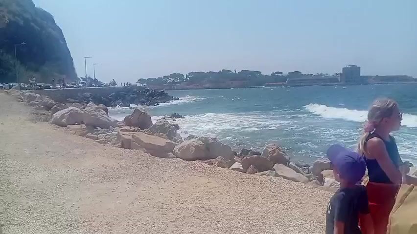 A group of people are walking along a rocky shoreline on a sunny day. A young boy wearing a purple hat is in the foreground, facing away from the camera. Behind him, a man and a woman are walking together, both carrying large bags. The woman appears to be wearing a pink shirt and the man is wearing a blue tank top. They are walking towards the water, which has waves crashing against the rocks. In the background, there is a hill covered in trees and some buildings. The sky is clear and blue.