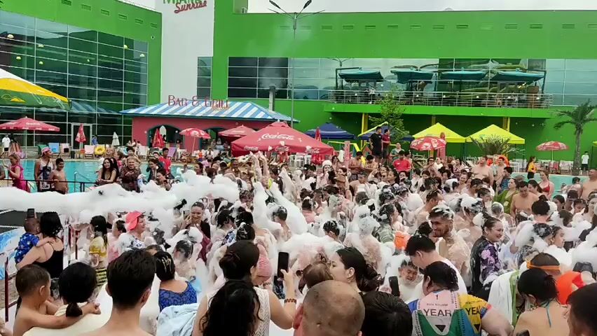 A foam party is taking place at an outdoor water park. A large crowd of people, including adults and children, are gathered in a pool filled with white foam. Some individuals are dancing and splashing in the foam, while others are watching the festivities. Various objects are visible in the background, including umbrellas, tents, and parts of buildings. The overall atmosphere appears to be festive and energetic.