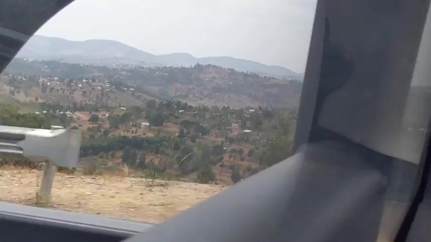 A view from inside a vehicle shows a hilly landscape with scattered buildings and trees. A metal guardrail runs along the side of a road in the foreground. A utility pole with some writing on it is visible to the right. The sky is overcast. The video appears to be recorded while the vehicle is in motion, as the scenery shifts slightly.