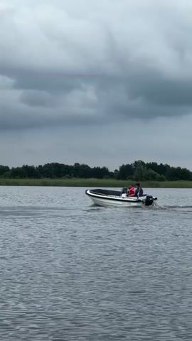 A small white motorboat with two people on board is moving from left to right across a body of water. The person in the front is wearing an orange life jacket and appears to be steering. The person in the back is wearing a dark jacket. A small outboard motor is attached to the back of the boat. The water is choppy, and there is a line of trees in the background under a cloudy sky. A Dutch flag is visible on the back of the boat.