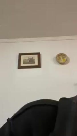 A man appears in the foreground, briefly looking at the camera. The background consists of a white wall with a picture and a small plate hanging on it. The ceiling is white and has a simple design. The man is wearing a dark-colored shirt.