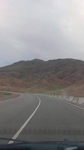 The video shows a view from the driver's seat of a car driving on a paved road. The road curves to the left, with a white line marking the center. The surrounding landscape is mountainous and arid, with reddish-brown hills covered in sparse vegetation. A concrete barrier runs along the right side of the road, with white posts marking the edge. The sky is overcast and appears to be a light gray. The video pans slightly to the left as the car navigates the curve.