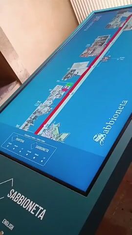 The video shows a close-up of a digital interactive map kiosk for Sabbioneta, Italy. The kiosk is a slanted rectangular screen mounted on a teal-colored base. The screen displays a map of the area with a red line running through it, and labelled points of interest. Text on the screen reads "Sabbioneta Select a video / select video" in both Italian and English. The kiosk has a touch screen interface.  A finger touches the screen near "Mantova." The kiosk is located indoors, next to a wall or partition with vertical lines. The floor is made of red brick.
