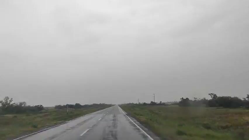 A view from inside a vehicle driving on a wet, paved road under a cloudy, overcast sky. The road is straight and stretches into the distance. On either side of the road are fields of grass and scattered trees. There are utility poles visible along the side of the road. The weather appears to be rainy, as indicated by the wet road surface and the overall grayness of the sky. No people or other vehicles are visible in the frame.
