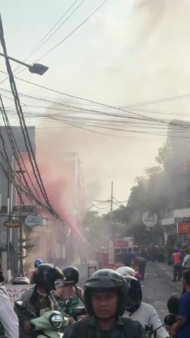 The scene captures a street in what appears to be a town or city, with a fire truck actively spraying a red liquid towards a building engulfed in thick, white smoke. Several onlookers, some wearing helmets and riding scooters, are observing the event from the foreground. There are buildings on both sides of the street, adorned with various signs and tangled overhead power lines. The overall atmosphere suggests an emergency response to a fire incident, with the focus on the firefighting efforts and the concern of the bystanders.