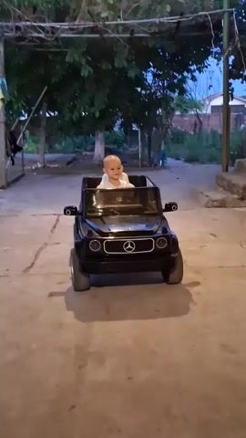 A bald baby sits in a black toy Mercedes G-wagon. The baby is smiling. A woman sits next to the car and appears to be holding a red object. The video is shot outdoors at dusk. There are trees and buildings in the background.