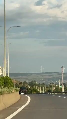 The video shows a view from a moving vehicle on a highway. The highway has multiple lanes and is bordered by a concrete barrier and lampposts on the left, and a metal fence and trees on the right. In the distance, there are rolling hills with wind turbines. Several cars are visible on the highway, including a black Jeep in the left lane and a white sedan further ahead. A tall cell tower stands prominently in the middle ground. The sky is partly cloudy with a mix of blue sky and grey clouds. The overall scene suggests a journey through a rural or semi-rural landscape under a dynamic sky.