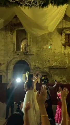 A bride throws a bouquet at her wedding reception while being held and spun by the groom. The reception is being held in a stone building, with a white sheet draped above the wedding party. Guests surround the couple, filming and cheering. There is a DJ booth in the back.