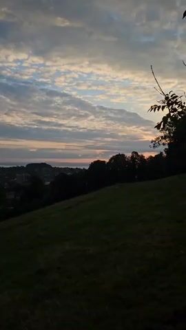 The video shows a view of a sunset over a town from a grassy hill. The sky is filled with clouds, and the sun is setting over the horizon, casting a warm glow. There are trees and buildings in the distance, and the hill in the foreground is covered in grass. The video is dark and grainy, suggesting it was filmed at dusk.