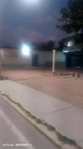 The video shows a street corner in what appears to be a small town, filmed at night. The camera is moving slightly, and the scene is dimly lit by streetlights. The sky is a dark blue with hints of purple. There are buildings on either side of the street, with closed shutters and doors suggesting that they are businesses or homes. A few trees are visible. A red car drives by in the distance. A tall, thin pole stands in the middle of the frame. The sidewalk is concrete. The quality of the video is low, with some blurriness. The bottom of the frame indicates the video was taken with an Infinix SMART 8.