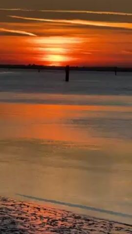 The video shows a sunset over a body of water. The sky is a mix of orange and red, with thin clouds creating a streaked effect. The sun is low on the horizon, and its light reflects on the water, creating a shimmering effect. There are a few poles sticking out of the water in the distance. The shoreline is visible in the foreground, with a dark, sandy texture. There are no people visible in the video. The scene is calm and peaceful.