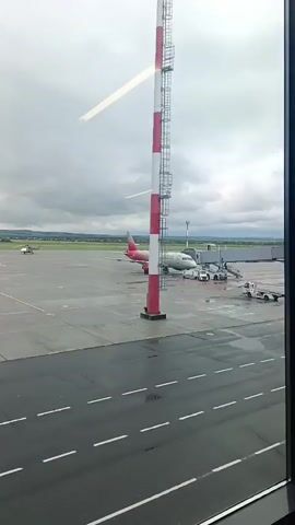 This media file shows an aerial view of an airport tarmac on a cloudy day. A commercial airplane with a red and white livery is parked at a gate, connected to a jet bridge. Several service vehicles, including a tug and a fire truck, are visible around the aircraft. In the distance, a smaller aircraft, possibly a helicopter, is seen on the runway. The tarmac is wet, indicating recent rain or damp conditions. The scene is framed by a window, suggesting the footage was taken from inside an airport terminal or a vehicle. The sky is overcast with dark clouds, and the overall atmosphere is muted and subdued. There are no people clearly visible in the frame, but the presence of the aircraft and service vehicles implies ongoing airport operations. The location is identified as Vyazovka, Russia. The time of day is indeterminate due to the overcast sky, but it appears to be daylight hours.