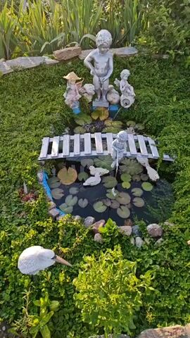 The video shows a small, decorative garden pond. The pond is surrounded by lush green ground cover and features several small garden statues. A miniature wooden bridge crosses over the pond. On the bridge, a statue of a child is depicted. In the pond, lily pads float on the water's surface, and there are two white duck statues. Behind the pond, at a higher elevation, a larger statue of a child stands. To the left of the larger statue, another small statue of a figure wearing a straw hat is visible. To the right of the larger statue, a statue of a seated child holding what appears to be a trumpet is present. In the foreground, a white statue resembling a swan or a stork lies on its side in the greenery. The scene is illuminated by natural sunlight, suggesting it is daytime.
