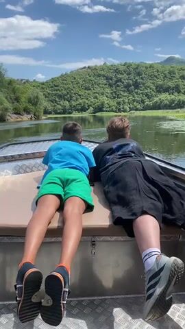 Two people, viewed from behind, are lying on the back of a boat. The person on the left is a child wearing a blue t-shirt and green shorts. The person on the right appears to be older, wearing a black shirt and dark pants. They are both looking out at a body of water surrounded by lush green hills under a partly cloudy sky. The boat is moving, as indicated by the rippling water and the shifting perspective of the shoreline. The overall scene depicts a leisurely boat ride in a scenic, natural environment.