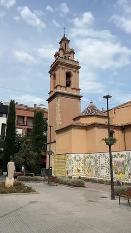 This media file features a view of a church with a prominent bell tower in Castelló De La Plana, Spain. The church has an orange facade and a tiled roof with blue accents. The bell tower is tall and has multiple levels, with bells visible inside. The surrounding area appears to be a public square or park, paved with patterned stones. There are benches, lampposts, and some greenery, including tall, slender trees and smaller bushes. A statue or monument is visible to the left of the church. Along the lower part of the church facade, there is a long, decorative tiled mural depicting various scenes and text. The sky is partly cloudy, suggesting it is daytime. The overall mood is peaceful and serene, capturing the architectural beauty of the church and its public setting. There are no people clearly visible in the frame. The visible text on the tiled mural appears to be in Spanish.
