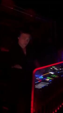 The video shows a DJ working behind a booth in a nightclub. The lighting is dark, dominated by red and some blue hues. There are two people behind the booth, one wearing headphones and manipulating the DJ equipment, and the other, partially obscured, standing to the side. The DJ booth has a red LED strip along its edge, highlighting the equipment, which includes turntables and a mixer with various buttons and controls. The environment is dark and indistinct, typical of a nightclub setting, with glimpses of other people in the background.