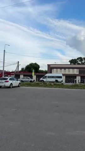A white van and a silver sedan are involved in a traffic accident. The van is facing right, and the sedan is facing left. The front of the sedan appears to be severely damaged. The vehicles are on a paved road, next to a grassy area and a building with a brown and tan facade. A pedestrian crossing sign is visible. The sky is cloudy.