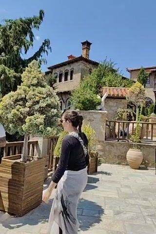 This video captures a scenic view of the Meteora monasteries in Kalambaka, Greece, on a bright, sunny day. The architecture of the monasteries, with their stone construction and domed roofs, is a prominent feature. Lush green trees and carefully maintained gardens surround the buildings, adding to the picturesque setting. Large terracotta pots are strategically placed throughout the courtyard, containing small trees and plants.

Several people are visible, appearing to be tourists exploring the site. They are dressed in casual, warm-weather clothing, suggesting a pleasant temperature. One person is seen walking across the paved courtyard, while others are gathered near the entrance to one of the buildings, possibly waiting to enter or admiring the view. The overall atmosphere seems relaxed and contemplative, with the clear blue sky and sunshine contributing to a serene mood. The paving stones of the courtyard are large and flat, laid out in an orderly pattern. The video pans across the scene, offering a panoramic perspective of the ancient structures and their natural surroundings.