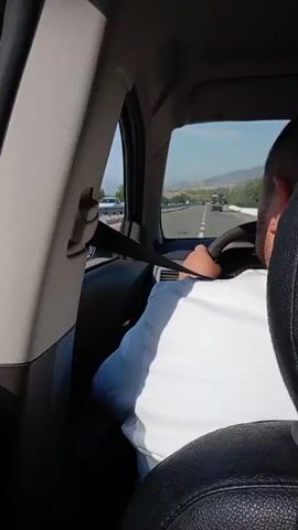 This media file captures a journey along a highway with a mountainous landscape in the background. The view is from inside a vehicle, looking out the window.

**Subjects:**
*   **Scene/Setting:** The scene is a highway with a metal guardrail separating it from a lower area. Beyond the guardrail, there is a sloping landscape with trees and distant mountains under a bright, sunny sky.
*   **Objects:** A highway guardrail, road markings (yellow lines), a car, and a white van are visible. The interior of the vehicle, including a window frame and a seat, is also in the foreground.
*   **People:** A person's arm and shoulder, wearing a blue garment, are visible in the lower right corner of the frame, indicating someone is a passenger in the vehicle. A person is partially visible inside the white car.

**Activity/Event:**
The primary activity is traveling by vehicle on a highway. The perspective suggests a moving camera, likely from a passenger's viewpoint. Other vehicles are also traveling on the highway, indicating ongoing traffic.

**Notable Details:**
*   **Time of Day:** The bright sunshine and clear sky suggest it is daytime, likely late morning or afternoon.
*   **Weather:** The weather appears to be clear and sunny.
*   **Location Cues:** The presence of a highway, guardrails, and the specific type of vegetation and mountainous terrain are consistent with a rural or semi-mountainous region. Gazalkent, Uzbekistan, is the provided location context.
*   **Emotions:** The visual information does not convey specific emotions. The scene suggests a typical travel experience.
*   **Visible Text:** No discernible text is visible in the media file.