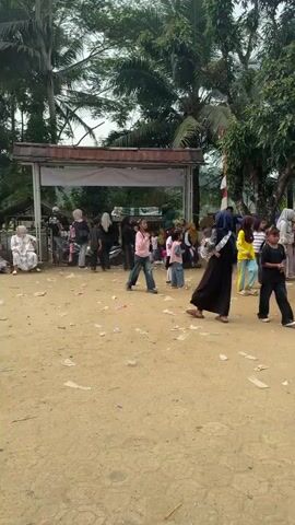 This video captures an outdoor gathering in Padamulya, Indonesia. A crowd of people, primarily children and adults, are milling around a makeshift stage area. The stage is covered by a white and blue tent, decorated with red, yellow, and purple balloons. Large speakers are set up on metal scaffolding to the right of the stage. 

The ground is dusty and appears to be a dirt or gravel area, with scattered pieces of paper or confetti. In the background, there are trees, including palm trees, and buildings, suggesting a rural or semi-rural setting. A few Indonesian flags are visible, indicating a possible national or local celebration.

The people are dressed in casual clothing, with many children in bright colors. Some individuals are wearing hijabs, common attire in Indonesia. The general atmosphere seems lively, with children actively moving around, some appearing to be dancing or playing. One child in the foreground is wearing a maroon shirt and blue shorts and is enthusiastically engaged in some activity. Another child in a yellow hijab and pants is also actively moving. The expressions on the faces of the children suggest excitement and enjoyment. The weather appears to be overcast, suggesting a possibly warm and humid day typical of tropical climates. The time of day is likely daytime, given the outdoor setting and the lighting.

The event seems to be a community gathering or festival, possibly with a performance or activity taking place on the stage. The presence of a stage, sound system, and a crowd suggests a planned event. No specific text is clearly legible in the video.