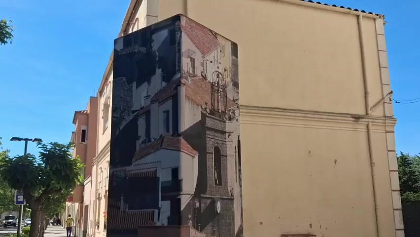 The video shows a street scene in La Jonquera, Spain. The camera pans down a tall, rectangular signpost with images and text related to the town. One side of the post features a black and white photo of the town's architecture, showing buildings with red-tiled roofs and a church tower. Below this is a depiction of people wearing traditional attire with plumes of smoke in the background. The third side of the post displays a map and text about La Jonquera, written in what appears to be the local language. The text describes the town’s history and mentions its location in Catalonia, near the border with France. A concrete bench sits to the right of the signpost. In the background, a street with sidewalks, trees, and buildings can be seen. A person wearing a yellow shirt is walking in the distance. The weather appears to be sunny with a clear blue sky.