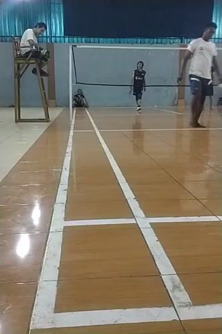 The video shows a badminton match played indoors on a wooden court. There are four players visible. One player is sitting on a metal chair on the left side of the net, holding a racket. On the other side of the net, there are three players in action. One player is in the air, attempting a shot, while another player is on the ground, possibly having just completed a shot or about to recover. A third player is also on the court, seemingly in a defensive position. The court is marked with white lines, indicating the boundaries of the playing area. The lighting appears to be artificial. The overall atmosphere suggests an active game of badminton.
