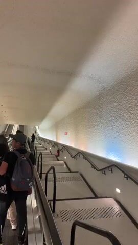 The video shows a group of people riding a long escalator upwards. The escalator has black handrails and white stairs with small black patterned dots. The walls and ceiling are off-white and slightly textured. The lighting is bright. Some people are wearing backpacks. An Adidas duffel bag is also visible. A red fire extinguisher is mounted on the wall near the top of the escalator. The escalator appears to be in a public space, possibly a transportation hub. The camera is facing upwards along the escalator.