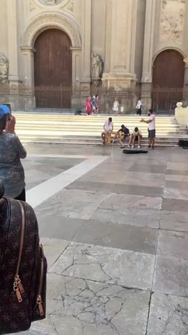 This video captures a lively street performance in front of a grand stone building, likely a cathedral given the architecture. Three young men are seated on steps, entertaining passersby with music. One plays a cajón, a box-shaped percussion instrument, while two others strum guitars. A fourth individual stands nearby, appearing to direct or participate in the performance, possibly singing or conducting.

Several people are visible on the steps and in the background, some pausing to watch the musicians, others continuing their day. The scene suggests a warm, sunny day, with the bright sunlight casting sharp shadows on the tiled plaza in the foreground. The setting is clearly outdoors, in a public square characterized by its ornate stonework and wide steps.

The overall mood appears upbeat and energetic, with the musicians focused and engaged in their performance, and the audience seemingly enjoying the impromptu concert. The cobblestone-like paving of the plaza, with its distinct geometric patterns, adds to the visual texture of the scene. The large wooden doors and the classical architectural elements of the building behind the performers point to a historical European location. There is no visible text within the frame of the video.