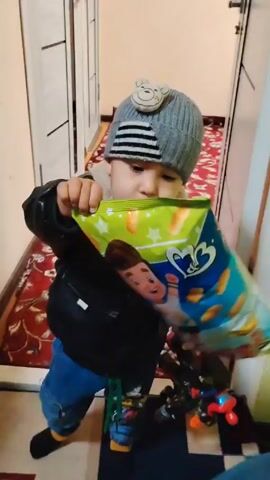 A young child in a gray knit cap, black jacket with faux fur collar, and blue jeans stands in a hallway with a red patterned rug. The child appears to be speaking while holding a toy in their hand. A cabinet with shelves is visible in the background. The video then goes dark.