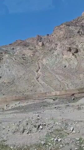 The video shows a mountainous landscape under a clear blue sky. The mountains are rocky and sparsely vegetated, with shades of gray and brown dominating the color scheme. A dirt road or path runs along the base of the mountain, separated by a low fence. The foreground is a rocky, barren area with scattered stones and a few patches of green vegetation. The video is taken from a moving vehicle.