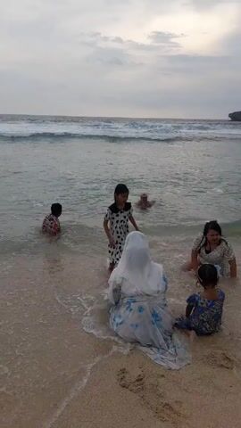 Several people are at the beach. The waves are coming in. There is a woman in a hijab sitting on the sand. Another woman stands in the water holding hands with a girl. A boy is sitting in the shallow water.  Someone is floating farther out in the water. The sky is overcast.