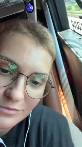 A close-up, slightly blurry video shows a person with short blonde hair and glasses looking into the camera. The person is wearing a black t-shirt and white earbuds. The background appears to be the interior of a bus, with a window showing a blurred landscape outside. The window has blinds partially covering it, and text is visible on the window, possibly a company name. The lighting suggests it is daytime.