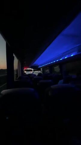 The video shows the interior of a bus at dusk or dawn. The bus is mostly empty, with only a few passengers visible in the distance. The overhead lights are blue. The camera then shifts to the window, revealing a view of the highway and a sunset. Cars and trucks pass by quickly.