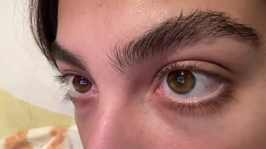 The video shows a close-up of a person's eyes. The eyes are brown, and the eyebrows are dark and thick. The skin is fair and slightly shiny. There is a faint reflection in the eyes. The background is out of focus, but appears to be a light-colored surface with some pattern.