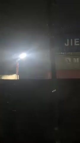 The video, shot at night, begins with a view over a dark wall toward a stack of shipping containers. A bright streetlight illuminates the scene, though the low lighting creates a high-contrast effect. The containers are labeled "JIEAN" and "MOL." As the camera angle shifts slightly, a tree comes into view, partially obscuring the container stack. Rain or snow is falling, visible as tiny white streaks in the air. The overall atmosphere is cold and industrial, with a sense of quiet activity taking place in the background.