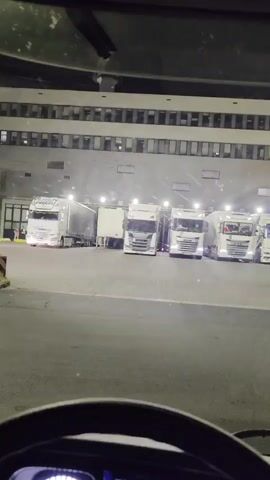 The video shows a view from inside a truck cab, looking out at a row of parked semi-trucks in front of a large building at night. The trucks are white and some have trailers. The building has many windows and appears to be a warehouse or distribution center. The lighting is artificial and casts bright spotlights on the trucks. The camera is slightly shaky, suggesting it is being held or mounted within a moving vehicle.