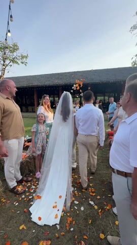 The video captures a joyful outdoor wedding procession or exit. A bride, wearing a long white wedding gown and a flowing veil, is walking away from the camera, holding hands with or walking closely beside the groom, who is dressed in a white long-sleeved shirt and light-colored trousers. They are walking down a grassy pathway, which is heavily covered with scattered orange and yellow flower petals.

Guests are lined up on both sides of the path, cheering and enthusiastically tossing more orange and yellow petals into the air and onto the couple as they pass. On the left side, a bald man in a light brown shirt and white pants stands smiling, watching the couple. Beside him, a young girl with distinctive blue-streaked braids and a floral dress is also observing. Further back on the left, a woman in a light top and patterned skirt is actively throwing petals. On the right side, several guests are visible, including a man in a white polo shirt and light pants watching the couple, and another guest holding up a phone to capture the moment.

The setting appears to be an outdoor venue, possibly a garden, with hay bales used as decorative elements lining the pathway. String lights with bare bulbs are strung overhead on the left side of the frame, hinting at an evening event or adding to the festive atmosphere. In the background, a large building with a dark, thatched or tiled roof and an open-air structure is visible. The sky is bright, suggesting late afternoon or early evening. The audio features sounds of cheering and exclamations like "Woohoo!", indicating a celebratory and festive mood among the guests.